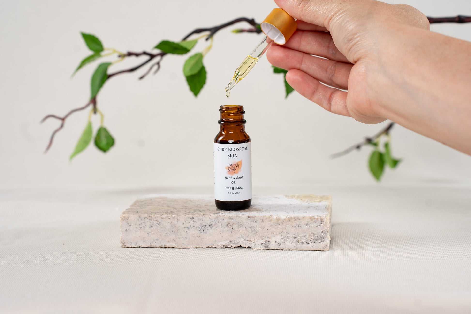 Hand holding gold trim dropper lid, with one drop of oil dropping into open 1/2oz amber glass bottle of Heal & Seal OIL | Pure Blossom Skin